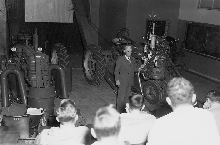 Harris P. Smith discusses cotton harvesting equipment in the lecture hall.  The tractors are on the wooden turntable that allowed rotation for better views by the students.
