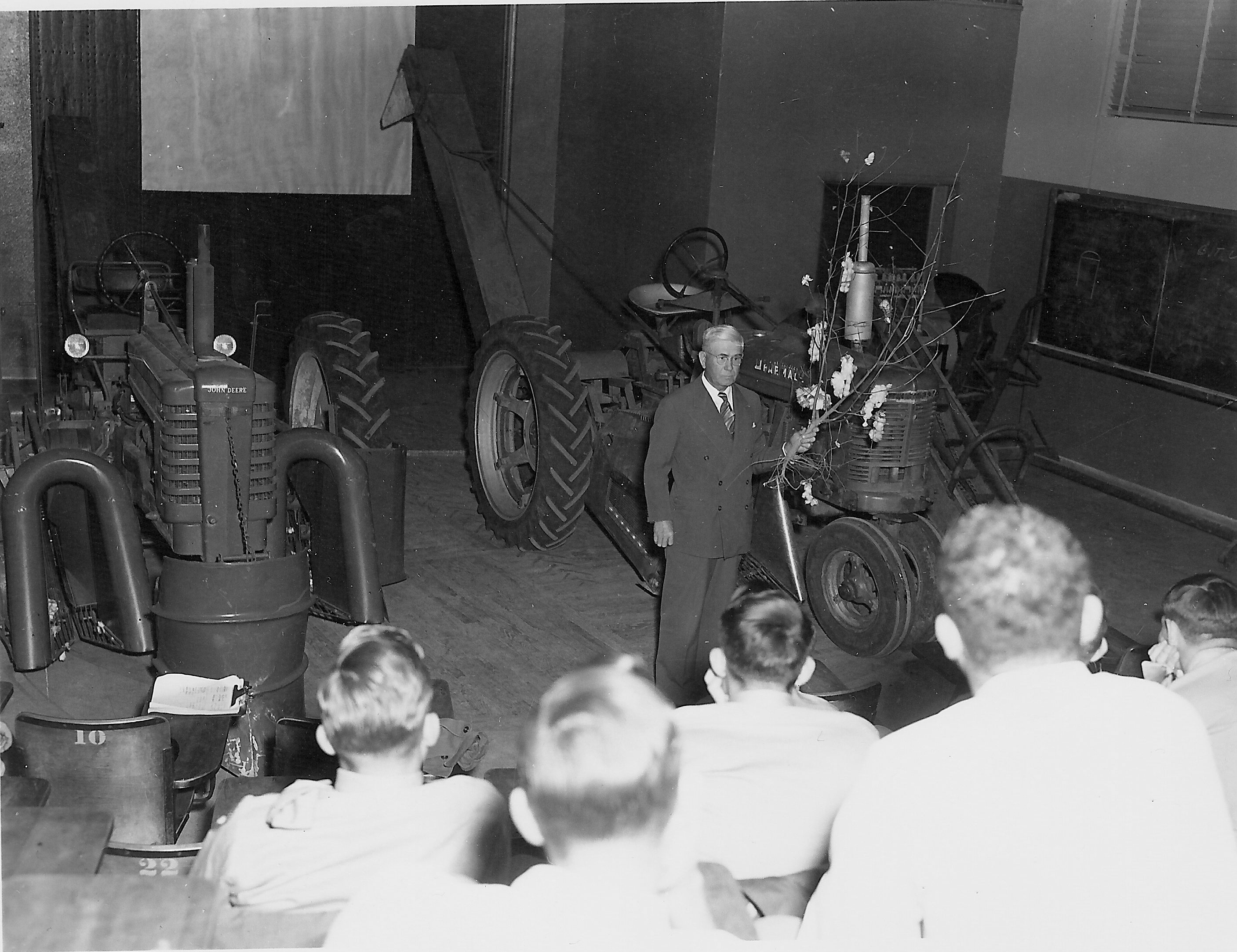 Harris P. Smith discusses cotton harvesting equipment in the lecture hall.  The tractors are on the wooden turntable that allowed rotation for better views by the students.