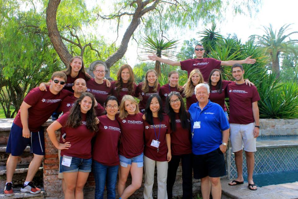 A group of students poses in maroon shirts with the founder of the program with Mexican palm trees in the background.